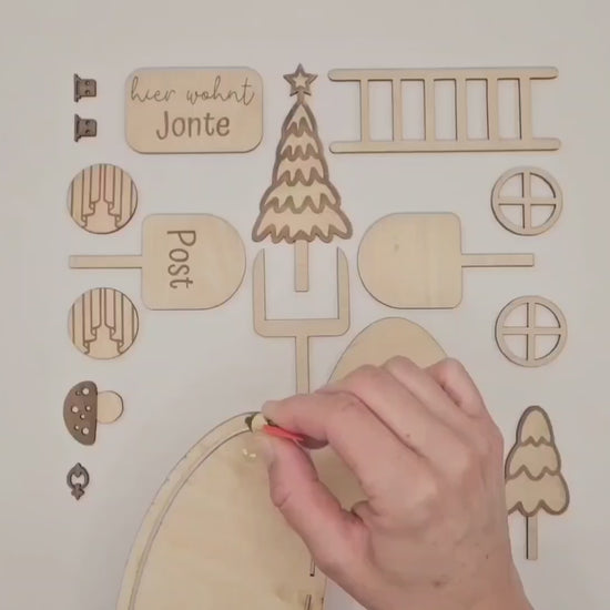 Personalisierbare Holz-Wichteltür mit Leiter, Briefkasten, Pilz und zwei Tannenbäumen an einer weißen Sockelleiste im Kinderzimmer, auf hellblauer Wand mit weißen Punkten montiert.